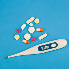 Pills and capsules of various shapes, digital thermometer on a blue background