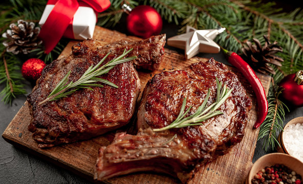 Christmas Dinner For Two, Grilled Beef Steak Ribeye, Herbs And Spices On A Stone Table With A Christmas Tree And New Year's Toys