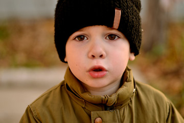 A small handsome boy in a back knitted hat and a brown jacket expresses an emotion of discontent 