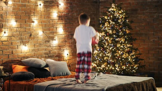 Cute little boy of 4 year old holding gift box and jumping on bed with excitement in cozy bedroom decorated with Christmas tree and garland