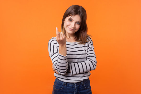Portrait of friendly pretty young woman with brown hair in long sleeve striped shirt standing, beckoning with finger to come nearer, smiling at camera. indoor studio shot isolated on orange background