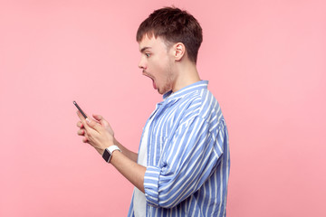 Side view of amazed brunette man with small beard and mustache in casual striped shirt holding phone, reading message with open mouth, astonished face. indoor studio shot isolated on pink background