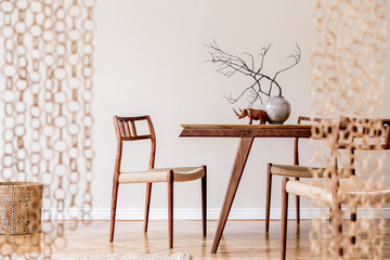 Stylish and beige interior of dining room with design wooden table and chairs, vase with flowers, sculpture, elegant and rattan accessories. Korean style of home decor.  Wooden parquet. Template.
