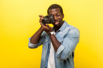 Portrait of passionate professional photographer, joyous man in denim casual shirt holding digital dslr camera and focusing, enjoying to take photo. indoor studio shot isolated on yellow background