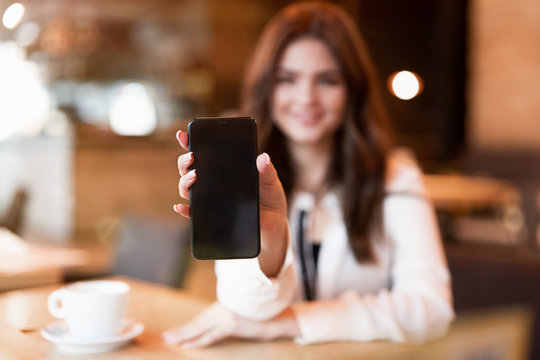 woman holding her smartphone showing device during lunch break drinking hot coffee in trendy cafe looking happy modern businesswoman