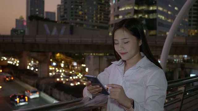 Side view of Young smiling business asian woman using smartphone in the modern city center in the night of Bangkok Thailand. Concept Technology communication by mobile phone