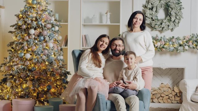 Cute European family of four posing on camera for a Christmas photo in the living room of his house in an armchair near the Christmas tree and fireplace. Speeding up the video at the beginning then