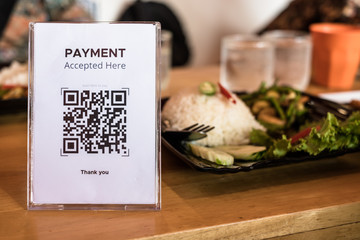 QR code payment accepted in acrylic or plexiglass table card holder on wooden table for restaurant with blurred food in the back. Mobile banking, commerce, finance technology.