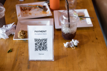 QR code payment accepted in acrylic table card holder on wooden table for restaurant with blurred leftover food after people ate in the back. Mobile banking, commerce, finance technology.