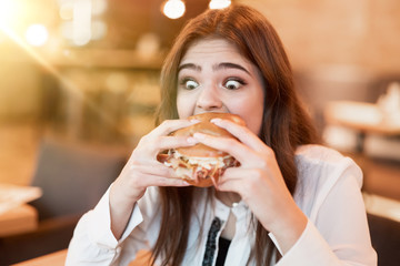 young woman in white stylish blouse biting with appetite fresh tasty meat burger during lunch in...