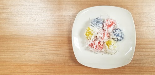 Flat lay or Top view of Colorful Khanom Tom Daeng (Coconut Balls) in white dish or plate on brown wooden table with left copy space. Dessert and Thai ancient food.