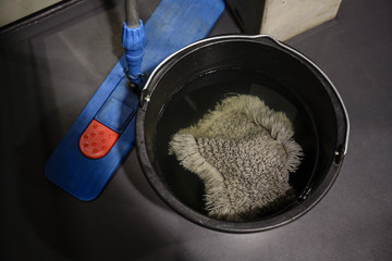 Wiping mop in a bucket with water and a flat handle for sweeping the floor on a dark surface in a modern building, work equipment for a low paid occupation in the cleaning industry