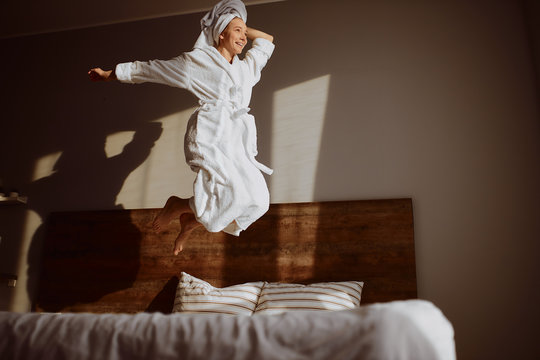 Beautiful joyful woman jumping happily on bed, looking away with cheerful expression, smiling brightly, raising leg high, having fun in bedroom, happiness lightness concept