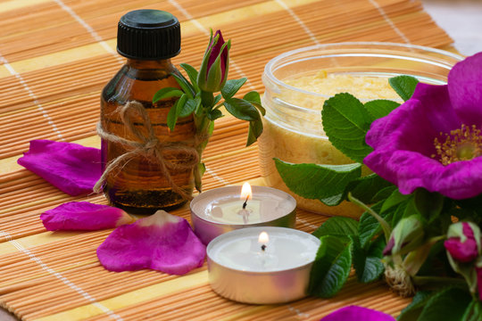 Dark bottle with rose bud with dog roses flowers petals oil nearby two candles and jar of sea salt with rose extract.