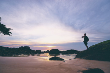 Silhouette of man stands on a rock at the beach while taking a photo of beautiful dusk using his phone.