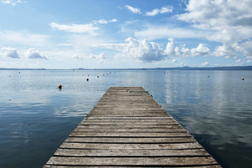 Wooden jetty for boats against a blue sky with some clouds. Floating buoys. Hills on background. Bolsena lake, Italy.