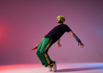 Attractive sportive man with thick beard, dressed in casual sportswear, having yellow cap and red bandana, standing on tiptoe, bending back with stretched hands, performing hard dance element