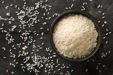 White uncooked, raw long grain rice in black bowl on rustic, black wooden table with copy space top...