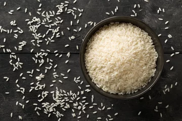 Fototapeten White uncooked, raw long grain rice in black bowl on rustic, black wooden table with copy space top view flat lay from above © Shawn Hempel