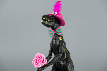 big plastic  dinosaur toy in hat, bow tie and with pink rose isolated on grey background, funny...