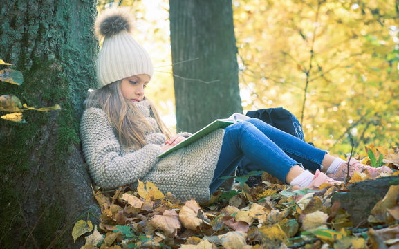 Cute little girl sitting with a book on her lap under the tree in public park. Girl sitting thoughtfully on the ground covered with yellow leaves in the autumn season sunny weather.