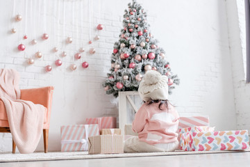 Little one year old girl in big warm hat plays with presented to her for new year in bright interior on background of Christmas tree and gifts in holiday cozy morning. White Home decoration New year