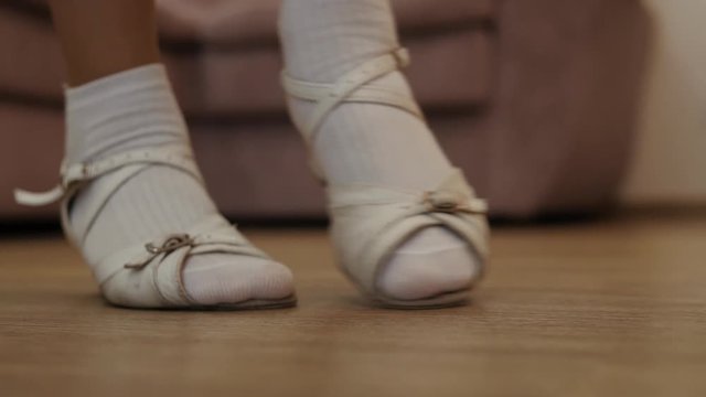 There are the feet of a girl taking dancing steps in a big living-room along the sofa. She is wearing white socks and sandals and moving like a ballerina. A great concept for a dance school.