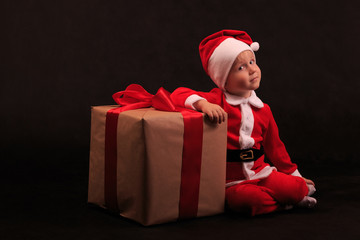 A child in a Santa Claus costume is sitting next to a gift tied with a red ribbon with a bow.