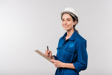 Happy young female builder in blue workwear and hardhat making notes in document