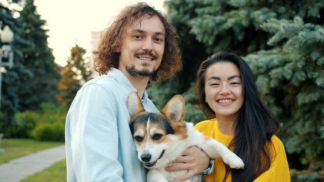 Portrait of young Caucasian man and Asian woman holding corgi puppy outdoors in city park smiling looking at camera. Lifestyle, people and love concept.