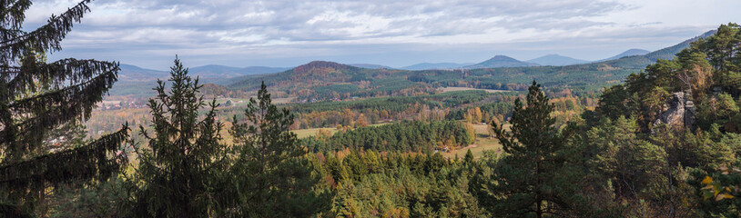 Panoramic view from Hrabencina vyhlidka on village Sloup v cechach in luzicke hory, Lusatian Mountains with autumn colored deciduous and coniferous tree forest and green hills, blue sky, white clouds