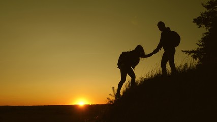 Obraz na płótnie Canvas male traveler holds the hand of a female traveler helping to climb top of the hill. Tourists climb the mountain at sunset, holding hands. team work of business partners. Happy family on vacation.