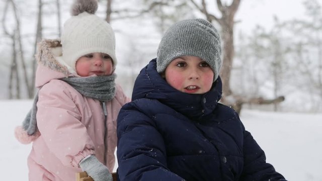 Daughter helps mother to pull sled with son on snowfall. Happy family sledding on snowy winter day. Cute little girl in jumpsuit pushes sled with her brother. Boy sledge outdoors on Christmas holidays
