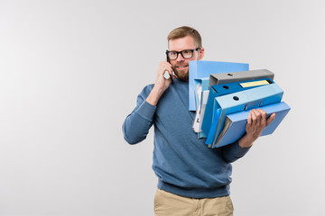 Busy young bearded manager with stack of folders holding smartphone by ear