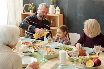 Grandfather pouring fruit juice into glass of his granddaughter by festive table