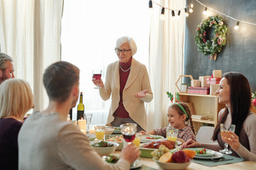 Senior grey-haired female toasting with glass of red wine in front of her family