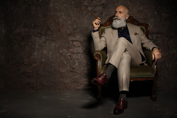 In chair sitting senior business man with cigar and whisky. Gray hair and beard wearing elegant...
