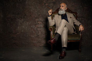 In chair sitting senior business man with cigar and whisky. Gray hair and beard wearing elegant suit. Against brown wall.