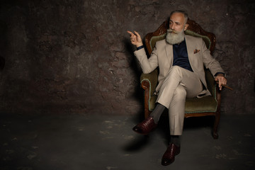 A portrait of a handsome mature man in a formal costume sitting in the armchair in the classic interior. Men's beauty, fashion.