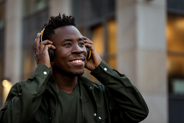 happy african-american man in stylish jacket in wireless headphones listening music on the street...