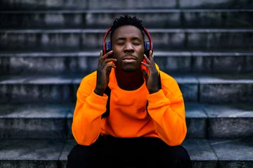 Foto op Aluminium african-american man in stylish orange hoodie sweatshirt in wireless headphones listening music and enjoying music on background of stairs © producer