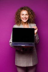 Portrait of a pretty smiling young blonde woman standing isolated over violet background, working on laptop computer. Young employee.