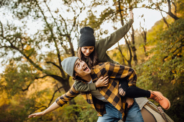 Happy loving couple of tourist have fun in the forest near tent and make airplane. Happy young man piggybacking his girlfriend while keeping arms outstretched.