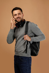 Fototapeta premium handsome young man with headphones and backpack talking on phone isolated on beige background.