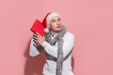 Man in a white sweater and red Santa hat shaking red box to find out what gifts are in.