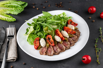 Food composition with Roast beef salad with sun-dried tomatoes. Arugula, tomato and cucumber on white plate on grey background close-up