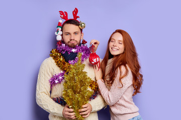 Pretty full of joy woman standing with closed eyes, smiling with pleasure, decorating bothered man with Christmas tinsel and baubles, having fun, pays no attention to angry male