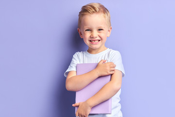 Portrait of blond kid boy holding book happily looking at camera , purple background. Kid involved in education