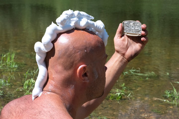 A bald guy with a shaving foam mohawk looks in the mirror. A man shaves in nature near a river with...