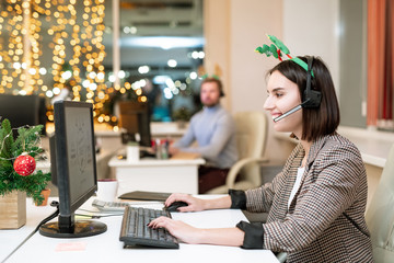 Happy young operator in xmas headband and headset shopping online by workplace